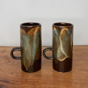 May include: Two tall ceramic mugs with a cylindrical shape and a small side handle. The mugs feature a dark brown base glaze with vertical, overlapping drips of tan and muted blue-green glaze that create a textured, layered appearance.