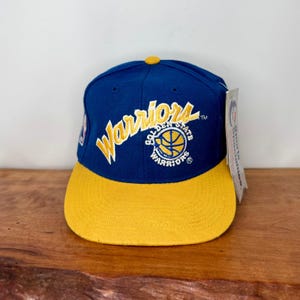 May include: A blue baseball cap with a yellow brim and a yellow button on top. The front features the word Warriors in yellow script and a circular Golden State Warriors logo. A white tag labeled Sports Specialties is attached to the side.