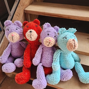 Puede incluir: Cuatro osos de peluche de ganchillo hechos a mano en tonos morado, rojo y azul. Cada oso tiene un hocico de color canela claro, ojos cosidos en negro y una nariz cosida en negro. Los osos están sentados en escalones de madera.