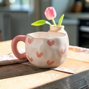 Puede incluir: Una taza de cerámica blanca con un borde irregular y ondulado y un asa rosa gruesa y redondeada. El exterior de la taza está decorado con pequeños corazones rosas pintados a mano dispersos por la superficie. La taza tiene un acabado brillante.