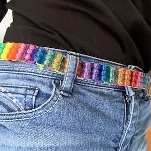 May include: A belt made of multi-colored translucent plastic pony beads arranged in a rainbow pattern, threaded onto a cord and worn through the belt loops of a pair of blue denim jeans with a black shirt tucked into the waistband.