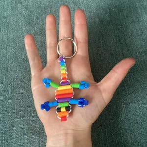 May include: A lizard-shaped keychain crafted from colorful plastic pony beads strung on a white cord. The lizard features a rainbow-colored body with blue, green, orange, red, purple, yellow, and black segments, attached to a silver metal ring.