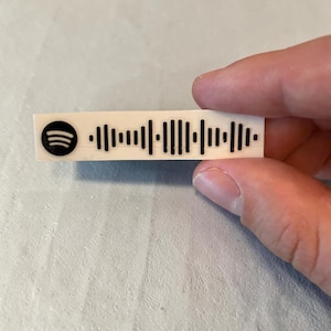 May include: A rectangular white sticker featuring a black Spotify logo on the left side followed by a black sound wave graphic. The sticker has a smooth finish and is designed to be applied to a surface for scanning purposes with a mobile device.