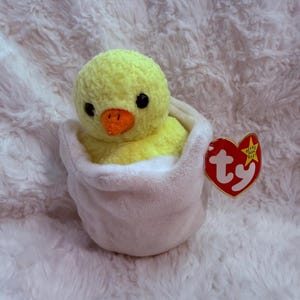 May include: A plush yellow chick with black eyes and an orange beak, nestled inside a white fabric egg shell. A heart-shaped red tag with the white text ty and a gold star is attached to the side of the egg, identifying it as a Beanie Baby.