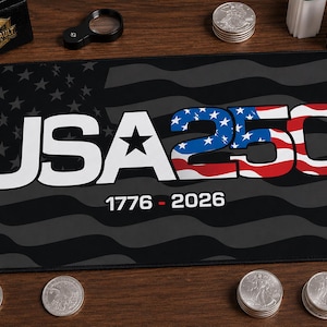May include: A rectangular black desk mat featuring a stylized American flag background in dark grey. The text USA250 is printed in large white and red, white, and blue letters, with 1776 - 2026 printed below. The Valley Vault Metals logo is in the corner.