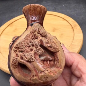 May include: A brown ceramic vase with a glossy neck and a matte, textured body featuring a relief carving of a pine tree and a small structure. The vessel has a rounded shape and a narrow opening, showcasing intricate details in the clay surface.