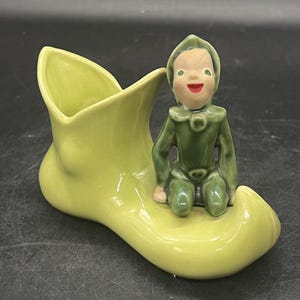 May include: A ceramic ornament showing a lime green boot-shaped vase with a seated elf figure. The elf is dressed in green with a smiling face and red lips. The boot has a smooth, glossy finish.