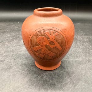 May include: A terracotta-colored ceramic vase with a round body and a narrow neck. The vase features a carved circular design with a figure holding a cross-like object. The vase is likely handmade and has a rustic, earthy aesthetic.