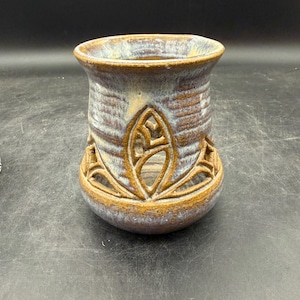 May include: A handcrafted ceramic vase with a unique openwork design. The vase features a cylindrical shape with a flared top and a detailed floral pattern carved into the body. The color palette includes shades of brown, blue, and tan.