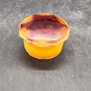 May include: A small, round, amber-coloured box with a dark brown lid. The lid has a scalloped edge and a swirl pattern.