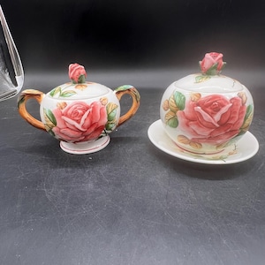 May include: A white ceramic sugar bowl and creamer set. The set features a large red rose design with green leaves. The creamer has brown handles. The sugar bowl has a lid with a rose bud on top. The set is on a black background.