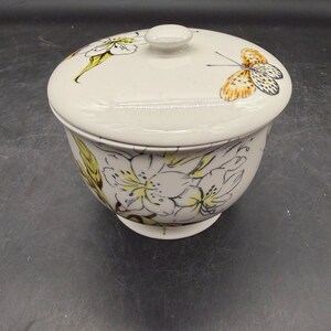 May include: Cream-colored ceramic bowl with a lid. The lid features a butterfly and floral design. The bowl is decorated with white flowers and green leaves. The bowl has a small, round knob on top.
