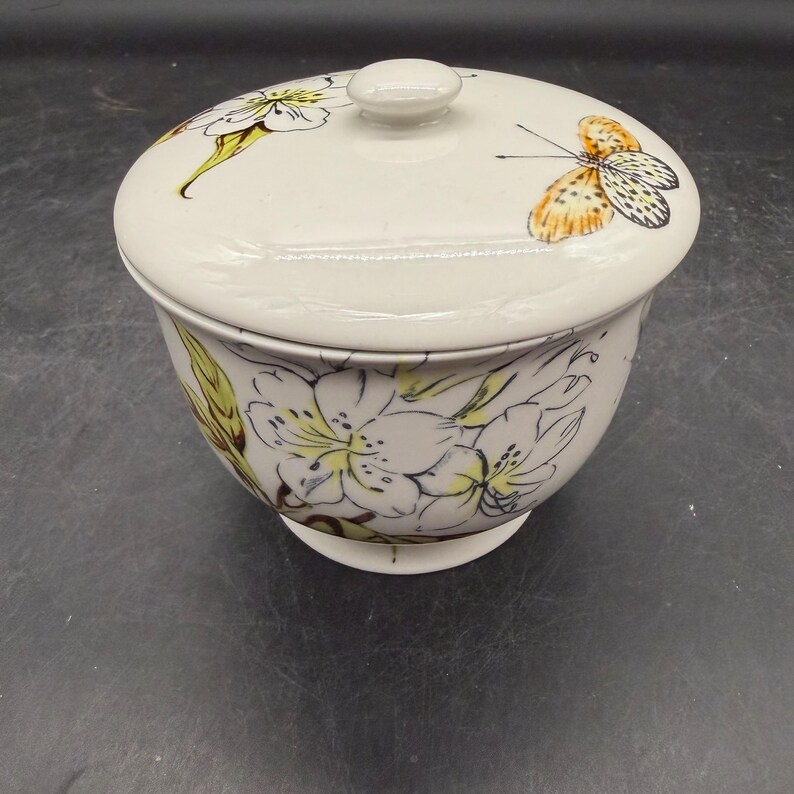 May include: Cream-colored ceramic bowl with a lid. The lid features a butterfly and floral design. The bowl is decorated with white flowers and green leaves. The bowl has a small, round knob on top.