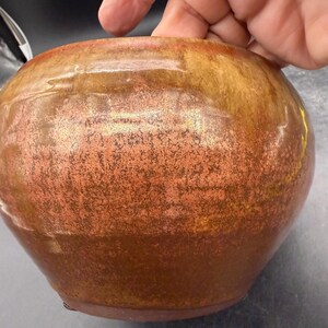 May include: A round, glazed ceramic bowl with a speckled copper and gold finish. The bowl has a wide, rounded body and a slightly flared rim. The surface has a textured appearance, with variations in color and tone.