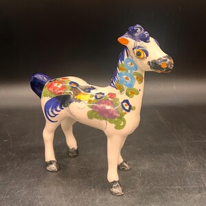 May include: A ceramic horse figurine with a cream-colored body, adorned with colorful floral patterns in red, blue, purple, and green. The horse has a blue mane and tail, and its hooves are painted gray. The figurine is standing on a dark surface.