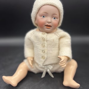May include: A vintage porcelain baby doll with blue eyes, rosy cheeks, and an open mouth. The doll is dressed in a cream-colored knitted hat, sweater, and shorts. The sweater has small, round buttons. The doll is sitting with its legs spread.
