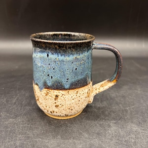 May include: Handmade ceramic mug with a speckled, two-tone design. The top half is a deep blue with dark speckles, transitioning to a speckled tan base. The handle is curved and matches the mug's color scheme.