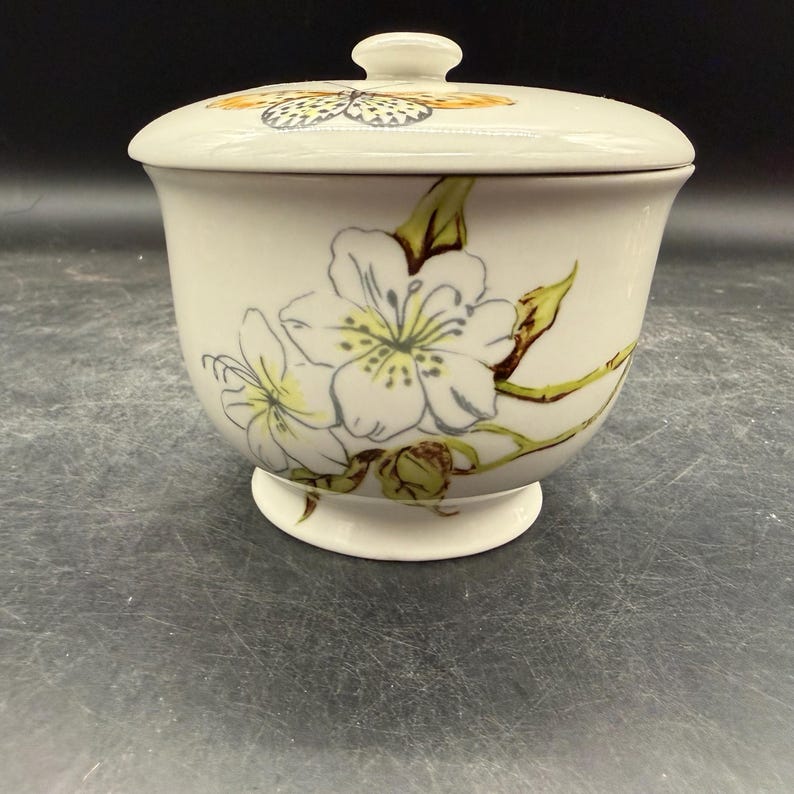 May include: Cream-colored ceramic jar with a lid, featuring a floral design and a butterfly on the lid. The jar has a pedestal base and is decorated with white flowers with yellow centers and green stems.