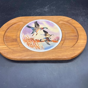 May include: Wooden serving tray with a central ceramic insert. The insert features a painting of ducks in flight over a lake with reeds and a pink tree. The tray has a light brown finish and a handle on each side.