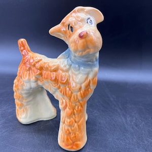 May include: A ceramic dog figurine with orange and white fur, blue accents, and a red nose. The dog is standing with its head turned to the side. The figurine has a glossy finish and is set against a dark background.