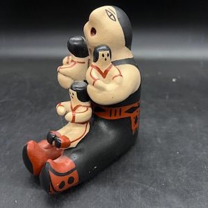 May include: A hand-painted ceramic figurine depicting a seated figure holding three smaller figures. The main figure is black with red accents, and the smaller figures are tan with red details. The figurine has a matte finish and is likely a collectible item.