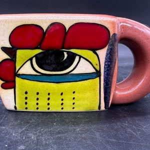 May include: A ceramic pitcher with a whimsical design featuring a large eye, red and green accents, and a brown handle.