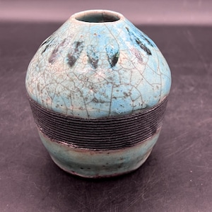 May include: A small, round, turquoise ceramic vase with a black band around the middle. The vase has a crackled glaze and a unique design.