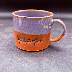 May include: A handmade ceramic mug with a lavender top and a reddish-orange base. The mug features the handwritten phrase "Made for More" on the base. The handle and interior are the same color as the base. A unique piece for coffee or tea.