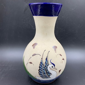 May include: A white ceramic vase with a blue rim and a painted design of a bird with blue and green wings. The vase has a round body and a narrow neck.