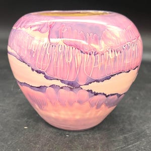 May include: A decorative, round ceramic vase with a glossy finish. The vase features a layered design of pink, lavender, and white hues. The colors blend and drip, creating a unique, artistic appearance. The vase has a wide opening and a rounded body.