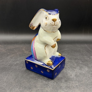 May include: A ceramic bunny figurine with a white body, blue hat, and gold accents. The bunny sits on a blue base decorated with gold floral designs. A rainbow stripe decorates the bunny's back.