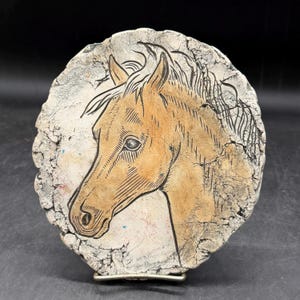 May include: A round, off-white ceramic piece with a horse head design. The horse is tan with black etched details, set against a textured background. The piece has a silver metal clasp on the back.