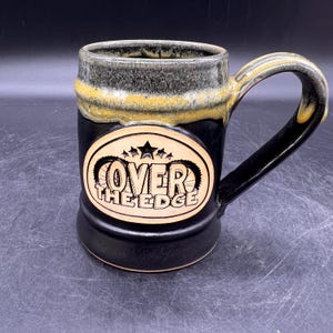 May include: Black ceramic mug with a yellow and gray glaze. The mug has a handle and a circular design on the front that says "Over The Edge" with stars and gears.