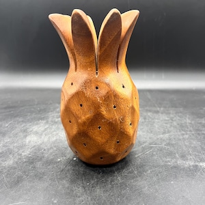 May include: A carved wooden pineapple with a brown finish. The pineapple has a textured surface with small holes and a crown of leaves at the top. The decorative item is set against a dark background.