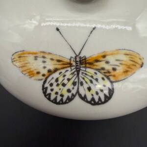 May include: A white ceramic lid featuring a detailed butterfly design. The butterfly has orange and yellow wings with black spots and a black body. The lid is round and has a small, rounded handle.
