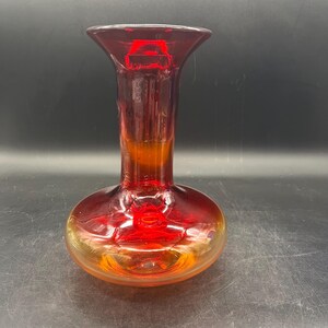 May include: A ruby red glass vase with a flared top and a rounded base. The vase has an ombre effect, transitioning from red to a golden hue near the base. The glass appears to be smooth and reflective.