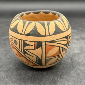 May include: A small, round, hand-painted ceramic pot with a wide opening. The pot features a tan base with black, orange, and gray geometric designs. The rim has a scalloped edge. The pot is approximately 4 inches in diameter.