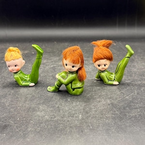 May include: Three ceramic figurines of elves in green outfits with red or blonde yarn hair. One elf is seated, while the other two are in yoga poses. The figurines have painted faces with rosy cheeks.