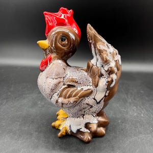 May include: A brown ceramic rooster figurine with a red comb and white speckles. The rooster is standing with its tail feathers raised.