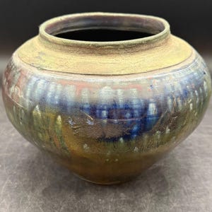 May include: A round ceramic vase with a wide opening and a textured, matte gold rim. The body features a multi-colored glaze with deep blue, green, and earthy brown tones, displaying a crackled finish and subtle vertical striations throughout.