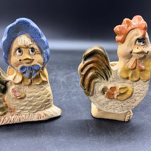 May include: Two ceramic figurines of chickens. One is a rooster with a brown, green, and yellow tail. The other is a hen wearing a blue bonnet.