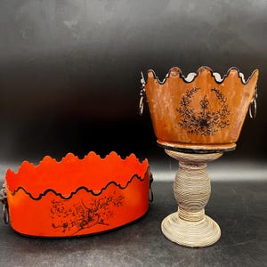 Peut inclure: Deux récipients décoratifs. L'un est un bol ovale orange vif avec un bord festonné et une bordure noire, avec une illustration noire. L'autre est une jardinière sur piédestal marron avec un motif floral et des poignées argentées.