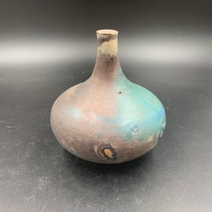 May include: A small, hand-thrown ceramic vase with a rounded body and a narrow neck. The vase is glazed in shades of blue, brown, and white, with a speckled finish.