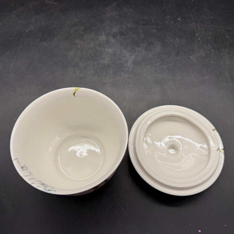 May include: Off-white ceramic container with a matching lid. The container has a rounded shape with a wide opening. The lid is circular with a small knob. Delicate floral and leaf designs in muted colors adorn the container's exterior.