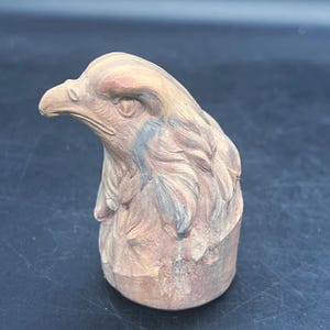 May include: A detailed sculpture of an eagle's head, showcasing intricate feather detailing and a sharp beak. The sculpture is primarily in shades of tan, with subtle hints of blue and gray, giving it a naturalistic appearance. The eagle's eye is clearly defined.