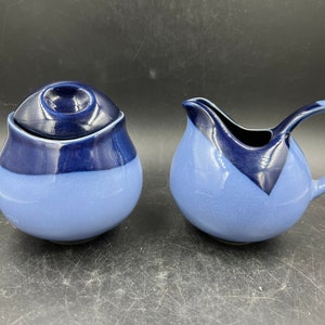 May include: A two-piece ceramic set featuring a sugar bowl with a lid and a creamer. The pieces are a combination of light and dark blue, with a speckled texture. The sugar bowl has a rounded shape, and the creamer has a handle and spout.