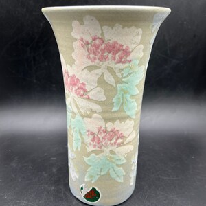 May include: A ceramic vase with a light brown background and a floral pattern in shades of pink, white, and green. The vase has a wide mouth and a narrow base.