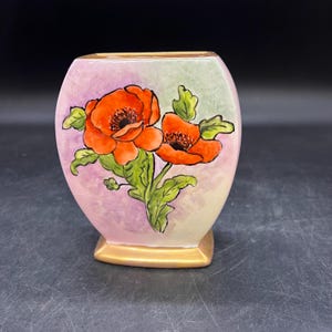 May include: A small, hand-painted ceramic vase with a square base and a rounded body. The vase features a floral design of two orange poppies with green leaves against a pastel pink and green background. The base is trimmed with gold.