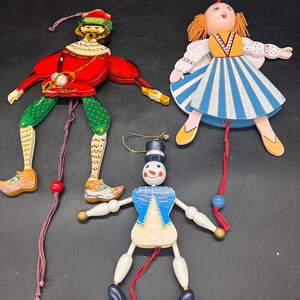 May include: Three wooden pull-string toys: a red and green jester, a doll with a blue and white striped dress, and a snowman with a blue top hat and jacket. Each toy has articulated limbs and is painted with bright colors.
