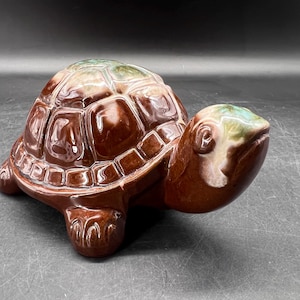 May include: A ceramic turtle figurine with a glossy finish. The turtle's shell is a rich brown with a geometric pattern, and the head and feet are a lighter brown. The top of the shell has green and white accents.